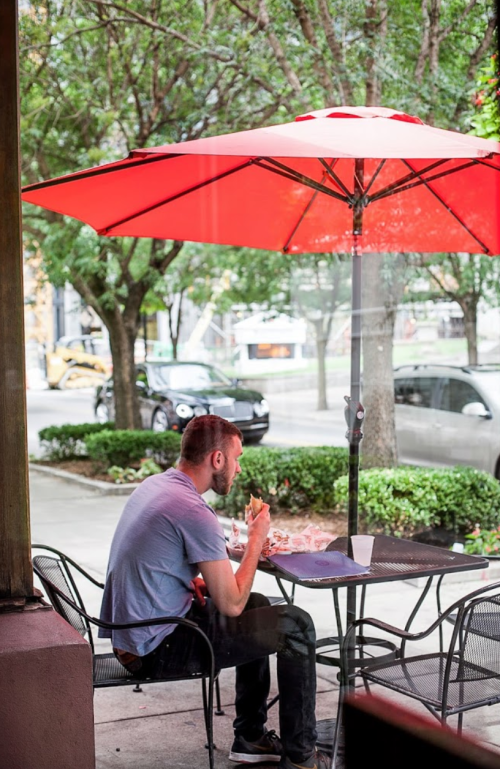 A man sits alone at a table under a red umbrella, enjoying a meal outside a café on a sunny day.
