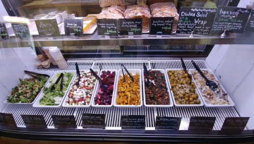 A display case filled with various salads and wraps, labeled with chalkboard signs, alongside baked goods.