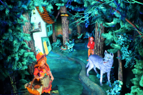 A whimsical forest scene with a house, a girl in a red hat, and a gray wolf among vibrant trees.