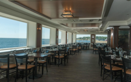 A spacious restaurant with wooden floors, tables set for dining, and large windows overlooking the ocean.