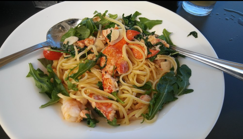 A plate of pasta with lobster, cherry tomatoes, and greens, garnished with herbs and served with two spoons.
