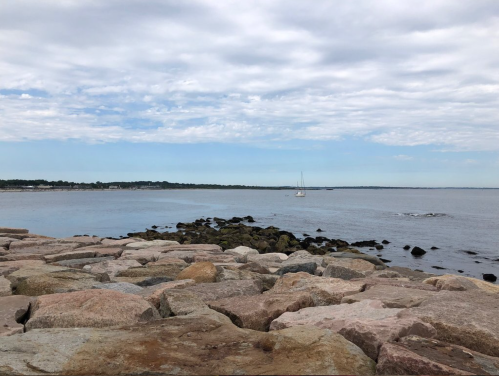 A rocky shoreline extends into calm waters under a cloudy sky, with a sailboat in the distance.