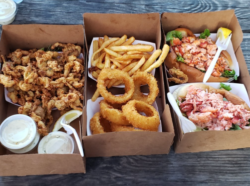 Three takeout boxes with fried seafood, including fried clams, onion rings, fries, and lobster rolls with lemon.