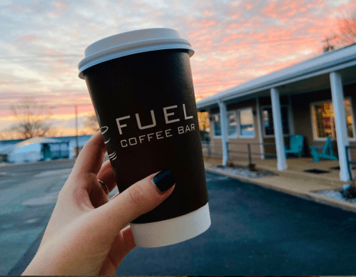 A hand holds a coffee cup with "FUEL COFFEE BAR" printed on it, against a colorful sunset backdrop.