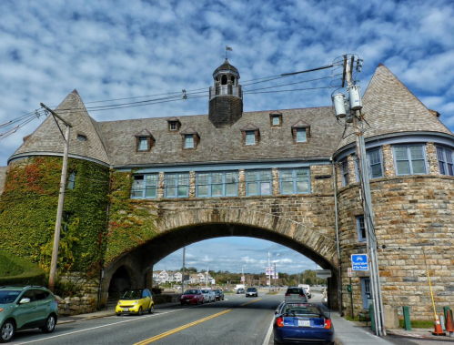 Historic stone building with a tower, archway, and ivy, set against a cloudy sky and surrounded by cars.