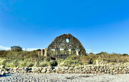 Ruins of a stone structure on a beach, surrounded by greenery under a clear blue sky.