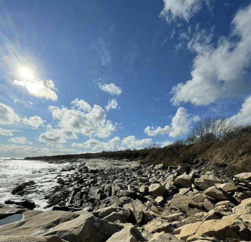 A rocky shoreline under a bright sky with fluffy clouds and the sun shining in the background.