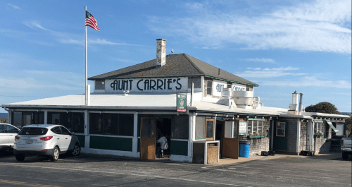 Aunt Carrie's restaurant with a flag, featuring a green and white exterior and outdoor seating. Clear blue sky above.