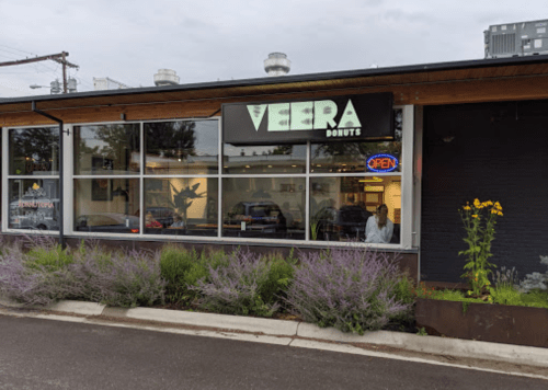Exterior of Veera Donuts with a large sign, "OPEN" neon light, and colorful flowers in front.