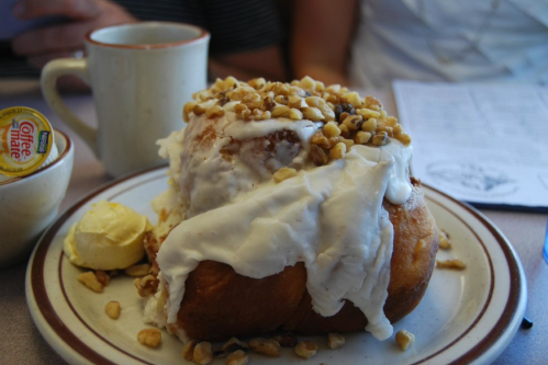 A large cinnamon roll topped with cream cheese frosting and chopped nuts, served with a pat of butter and coffee.