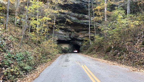 A car drives through a rocky tunnel surrounded by autumn foliage along a narrow road.