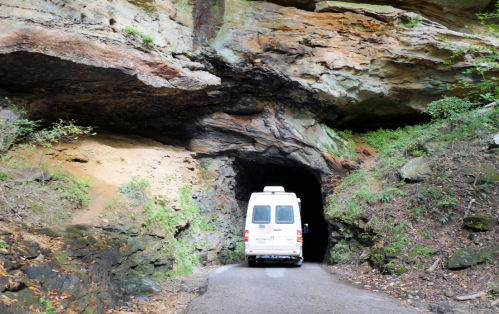 A white van drives through a rocky tunnel surrounded by lush greenery and rugged cliffs.