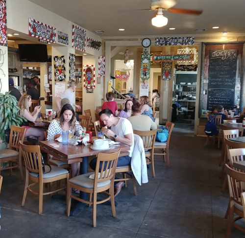 A busy restaurant with diners at wooden tables, colorful wall art, and a chalkboard menu in the background.