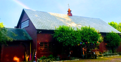 A red barn-style building with a slate roof, surrounded by greenery and colorful flags, under a clear blue sky.