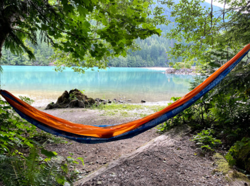A vibrant orange hammock hangs between trees by a tranquil lake, surrounded by lush greenery and mountains in the background.
