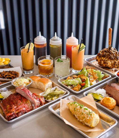 A spread of barbecue dishes, sides, and drinks on a table, featuring ribs, coleslaw, corn, and various sauces.