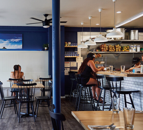 A cozy restaurant interior with wooden tables, black chairs, and patrons at the counter enjoying their meals.