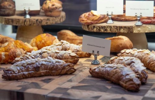 A display of various pastries, including almond croissants, muffins, and sweet rolls on wooden stands.