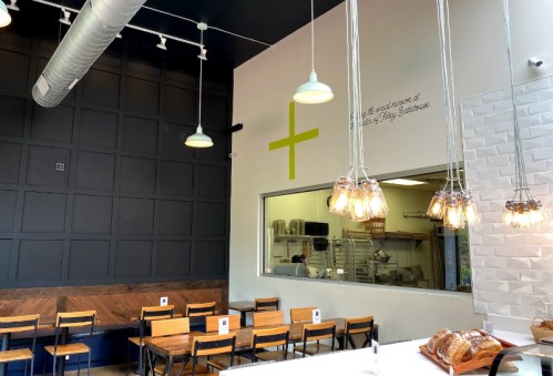 A modern café interior with wooden tables, pendant lights, and a large green plus sign on the wall.