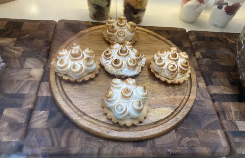 A wooden platter displays six decorative meringue pastries with swirled tops, arranged in a circular pattern.