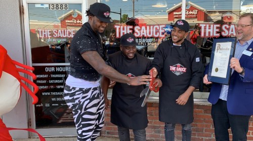 Three men in black aprons cut a ribbon outside a restaurant called "The Sauce," celebrating its opening.