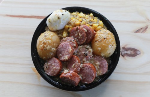 A black bowl filled with sausage, potatoes, corn, and a poached egg, served on a wooden surface.