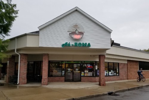 Exterior of Ah-Roma restaurant with a sign and open neon light, featuring a brick facade and a cyclist passing by.