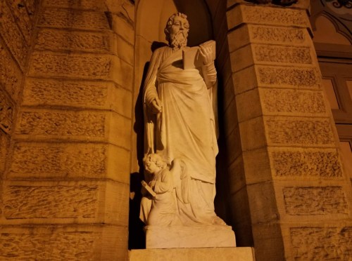 A white statue of a bearded man holding a book, with an angelic figure kneeling beside him, illuminated at night.
