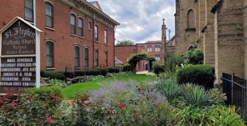 A well-maintained garden with flowers and greenery beside St. Stephen Catholic Church, featuring a sign with service times.