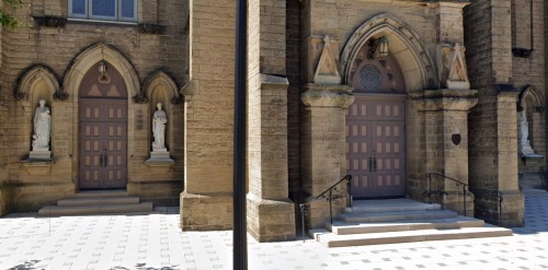 Two ornate church doors with statues on either side, framed by stone arches and a patterned walkway.