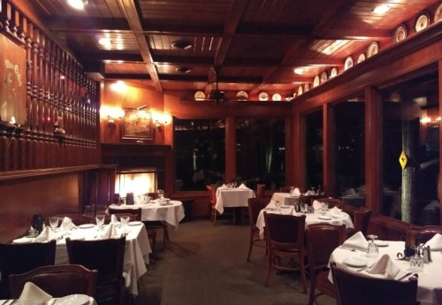Cozy restaurant interior with wooden decor, tables set with white linens, and a fireplace in a warm, inviting atmosphere.