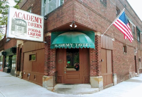 A brick building with a green awning and an American flag, featuring a sign for Academy Tavern, a food and liquor establishment.