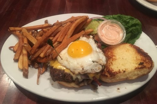 A juicy cheeseburger topped with a fried egg, served with sweet potato fries and a side of dipping sauce.