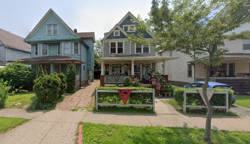 Two houses side by side, one blue and one light green, with a porch and a red Superman symbol on the fence.