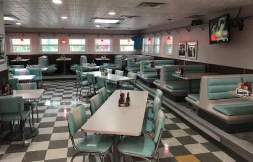 A retro diner interior with teal booths, checkered floor, and tables, featuring ketchup and mustard bottles on each table.