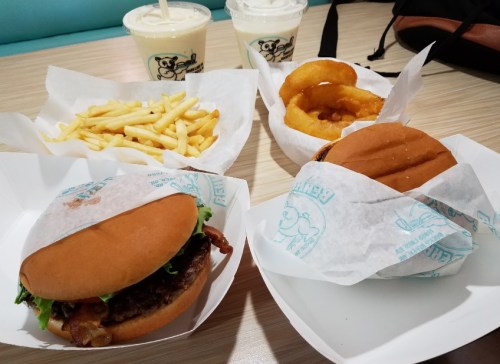 A table with two burgers, onion rings, fries, and two milkshakes in paper containers.