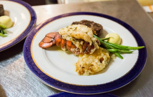 A gourmet plate featuring lobster, shrimp, steak, green beans, and creamy mashed potatoes on a decorative dish.