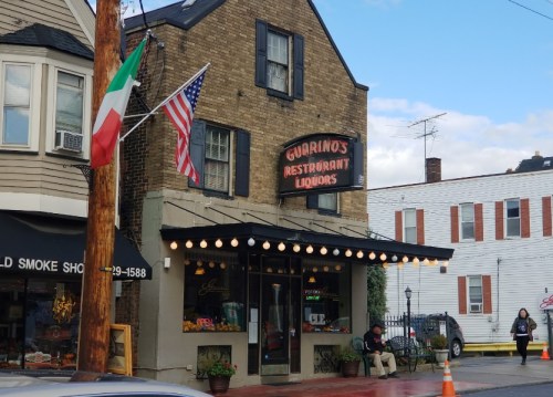 A brick building with a sign reading "Guarino's Restaurant Liquors," featuring flags and outdoor seating.