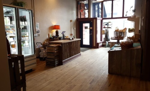 A cozy shop interior with wooden floors, a counter, a fridge, and a welcoming entrance filled with natural light.