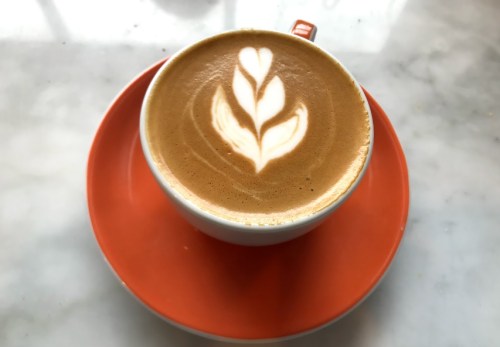 A cup of coffee with a heart-shaped latte art design, sitting on an orange saucer against a marble background.