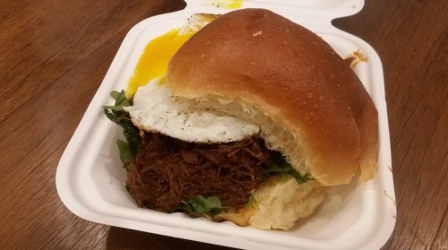 A pulled beef sandwich with a fried egg and greens, served in a takeout container.