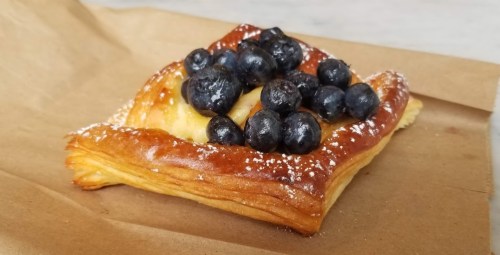 A flaky pastry topped with fresh blueberries and a dusting of powdered sugar, resting on brown paper.