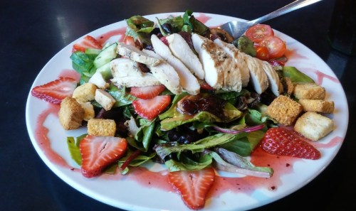 A colorful salad topped with sliced grilled chicken, strawberries, cucumbers, tomatoes, and croutons on a white plate.