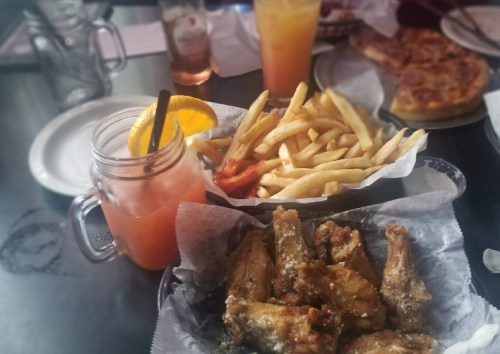 A table with a drink, fries, and a plate of chicken wings, with pizza in the background.