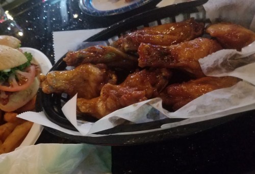 A basket of glazed chicken wings on parchment paper, with a side of sandwiches and fries in the background.
