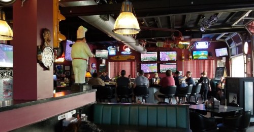A lively bar scene with patrons seated at the counter, multiple TVs showing sports, and colorful neon signs.