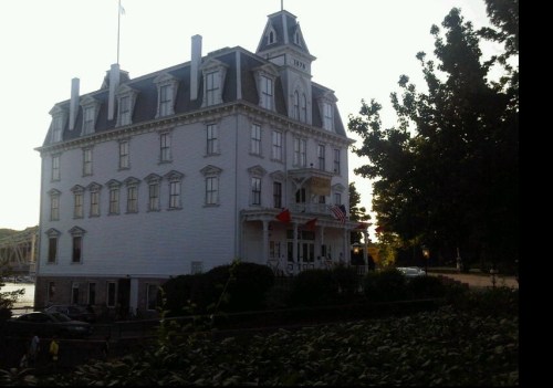 Historic white building with a peaked roof, surrounded by trees, flags flying, and a clear sky in the background.