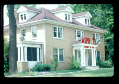 A large, two-story brick house with a red sign reading "WIPB" in front, surrounded by trees and greenery.