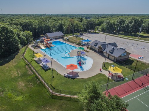Aerial view of a swimming pool with slides, surrounded by green trees and a tennis court nearby.