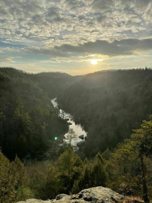 A scenic view of a river winding through a valley, surrounded by trees, with a sunset illuminating the sky.